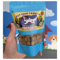 A hand holding a blue stand-up pouch of dog treats. The front label reads 'PUT YOUR PAWS UP AND CHILL' with a cartoon dog on a couch, and a clear window shows the brown flower shaped treats inside. The label states that the treats are made with chamomile, lavender and lemon balm, and that they are grain-free and hypoallergenic.