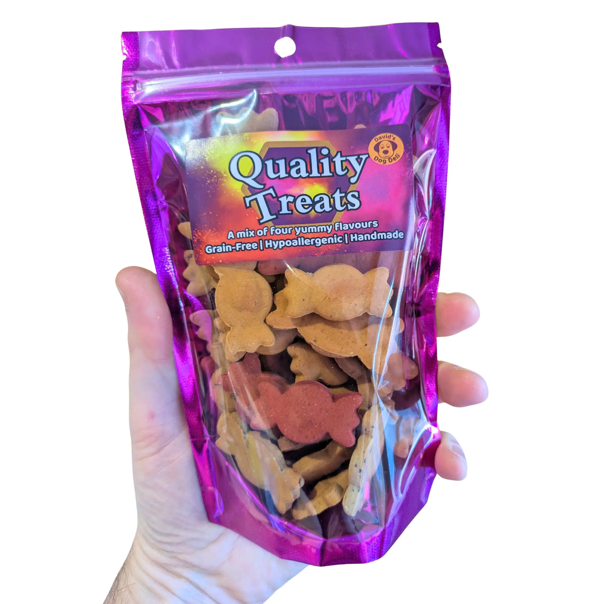 A hand holding a clear plastic bag of dog treats with a shiny purple/magenta stand-up pouch. The front label reads 'Quality Treats' and shows several sweetie-shaped dog treats visible through the clear window.