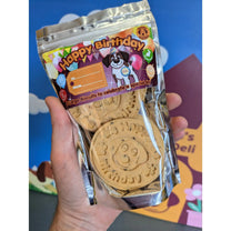 A hand holding a clear plastic bag of dog birthday biscuits. The packaging features a colorful 'Happy Birthday' label with a cartoon dog and a blank 'To: / Name:' field.