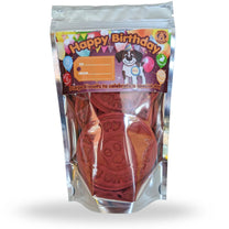 A stand-up pouch of personalized birthday dog treats, featuring a colorful 'Happy Birthday' header card with a dog illustration and a clear window showing round red-brown stamped biscuits inside.