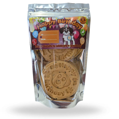 A package of dog birthday biscuits in a clear stand-up pouch with a colorful 'Happy Birthday' label. Two round tan biscuits are visible inside, embossed with 'Happy Birthday' and a dog face.