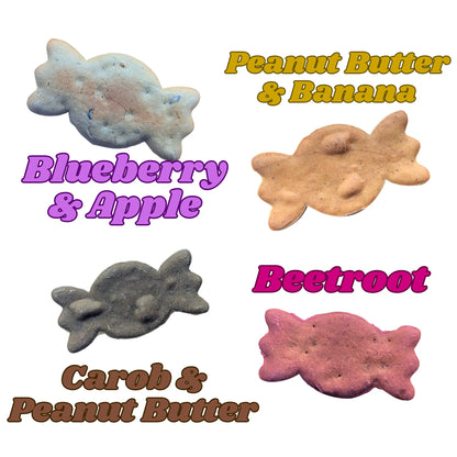 Four sweetie shaped dog treats arranged on a white background with colorful text labels: Blueberry & Apple, Peanut Butter & Banana, Carob & Peanut Butter, and Beetroot.