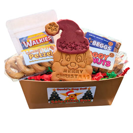A Christmas-themed dog biscuits selection box from David's Dog Deli (dog treat bakery), featuring a large embossed dog-shaped cookie wearing a maroon Santa hat with 'MERRY CHRISTMAS' written on it, surrounded by assorted dog treats including Doggy Donuts, Beggs, Walkies and Cheesy Petzels in a gold ballotin box with a branded label.